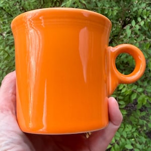 Puede incluir: Una taza de cerámica blanca con un asa negra y el texto "But First, Coffee" en un estilo manuscrito. La taza está sobre una superficie blanca. La taza mide 10 cm de alto.