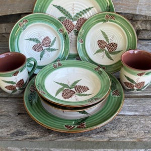 May include: A rustic dinnerware set with plates, bowls, and mugs. The set has a cream-coloured centre with a brown pinecone and green leaf design. The rims are green with a textured pattern. The mugs have a brown interior.