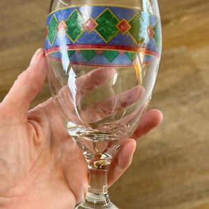 May include: A clear wine glass with a decorative band featuring green, red, and blue geometric patterns. The glass has a stem and a round base. The glass is held by a hand.