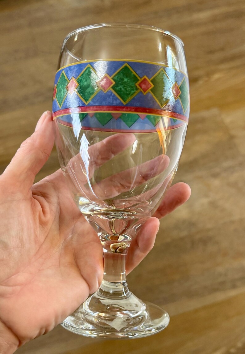 May include: A clear wine glass with a decorative band featuring green, red, and blue geometric patterns. The glass has a stem and a round base. The glass is held by a hand.