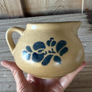 Può includere: Una brocca in ceramica beige con corpo arrotondato e manico. La brocca presenta un motivo floreale blu e un beccuccio per versare. La brocca è tenuta nella mano di una persona.
