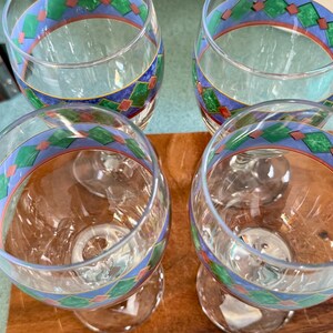 May include: Four clear wine glasses with a decorative band featuring blue, green, and red geometric patterns. The glasses are on a wooden surface, showcasing their elegant design and potential for serving beverages.