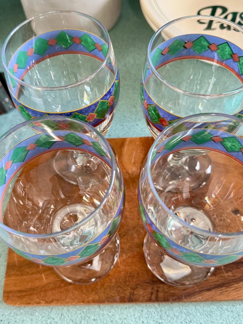 May include: Four clear wine glasses with a decorative band featuring blue, green, and red geometric patterns. The glasses are on a wooden surface, showcasing their elegant design and potential for serving beverages.