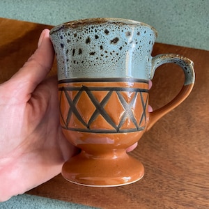 以下が含まれることがあります： 青と茶色の釉薬がかかった茶色の陶器のマグカップ。マグカップは中央に幾何学模様があり、広がった底になっています。