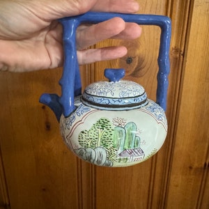 Può includere: Una piccola teiera blu e bianca con un design di paesaggio dipinto. La teiera ha un manico a forma di ramo.