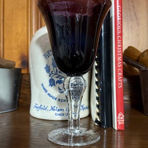 May include: A vintage wine glass with a deep purple bowl and a clear, ornate stem. The glass sits on a round base. The background includes a ceramic jug and a book with a red spine.