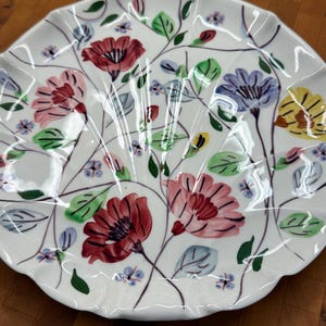 May include: White ceramic serving plate with a scalloped edge, adorned with a floral pattern. The design features red, pink, purple, and yellow flowers, along with green leaves and blue accents. The plate has a delicate, hand-painted appearance.