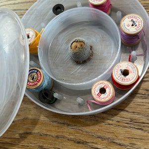 May include: A clear plastic sewing thread organizer with a variety of spools of thread in different colors, including pink, red, blue, and yellow. The spools are labeled "Coats & Clarks" and have different thread sizes.