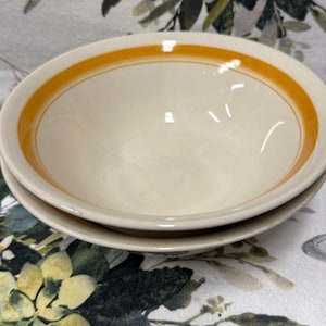 Może przedstawiać: Dwie kremowe ceramiczne miski ułożone jedna na drugiej. Każda miska ma cienki, pomarańczowy pasek wokół krawędzi. Miski są ustawione na powierzchni z kwiatowym wzorem.