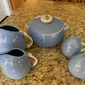 Może przedstawiać: Zestaw vintage niebieskich i białych ceramicznych naczyń kuchennych. Zestaw zawiera pokrywkę do zapiekanek, dwa dzbanki do śmietanki i dwie solniczki i pieprzniczki. Elementy zdobi kwiatowy wzór z białym obramowaniem.