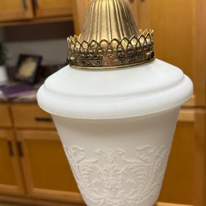 May include: Vintage pendant light with a white, bell-shaped shade featuring an embossed floral design. The light fixture has a gold-colored metal crown and top, and a chain for hanging.