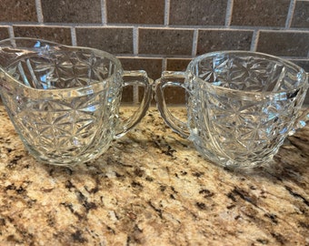 Op de afbeelding: Een vintage set van een roomkan en suikerpot van glas met een helder, ruitvormig patroon. De set is gemaakt van helder glas en heeft een delicaat, elegant ontwerp.