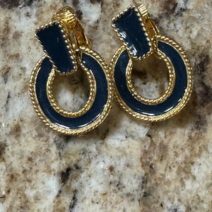 May include: A pair of gold-tone clip-on earrings with black enamel. The earrings are shaped like a ring with a smaller black enamel square in the center.