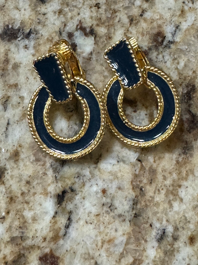 May include: A pair of gold-tone clip-on earrings with black enamel. The earrings are shaped like a ring with a smaller black enamel square in the center.
