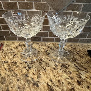 May include: Two clear crystal stemmed glasses with intricate cut designs. The glasses are on a brown speckled countertop.