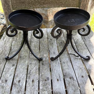 May include: Two black metal candle holders with a round top and three curved legs. The candle holders have decorative scrollwork and are placed on a weathered wooden surface. The candle holders are designed to hold candles.