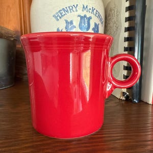 Puede incluir: Una taza de cerámica de color rojo brillante con un acabado liso y lustroso y un asa circular. La taza presenta un cuerpo cilíndrico sencillo con un sutil detalle de anillo horizontal cerca del borde superior, ofreciendo un diseño funcional.