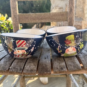 May include: Four navy blue ceramic bowls with a snowman design. Each bowl has a white interior and a dark blue exterior decorated with a snowman wearing a hat and scarf, with snowflakes and Christmas tree accents. The bowls are placed on a wooden surface.