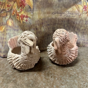 May include: Two terracotta turkey-shaped planters. The planters are light brown with carved details, including feathers and facial features. They are set against a backdrop of a fall-themed fabric with a signature.