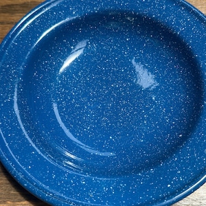 May include: A round, deep bowl with a glossy finish, featuring a dark blue base color and a speckled white pattern throughout the surface. The bowl has a wide, flat rim and a smooth, reflective texture, showcasing a classic enamelware design.
