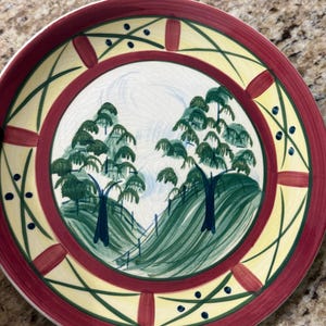 May include: A round ceramic plate featuring a hand-painted landscape scene with two green trees on rolling hills. The border is decorated with a wide maroon band, yellow sections, and thin green lines with small dark blue dots throughout the design.