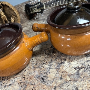 Może przedstawiać: Dwa brązowe garnki ceramiczne z pokrywkami i uchwytami. Garnki mają ciemnobrązową glazurę na górze i jasnobrązową glazurę na dole.