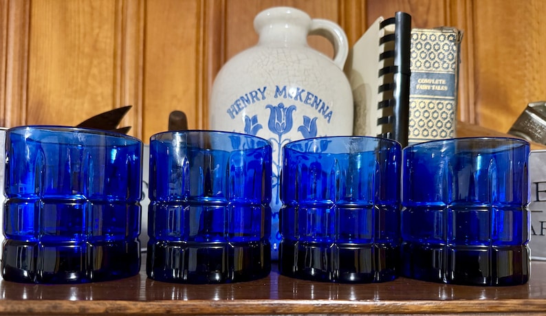 May include: Four cobalt blue glass tumblers with a square-shaped design. The tumblers are arranged in a row on a wooden surface.