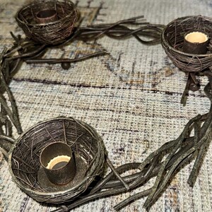 May include: A rustic metal candle holder with three candle cups shaped like bird nests. The holder is made of twisted metal branches and has a dark brown finish.