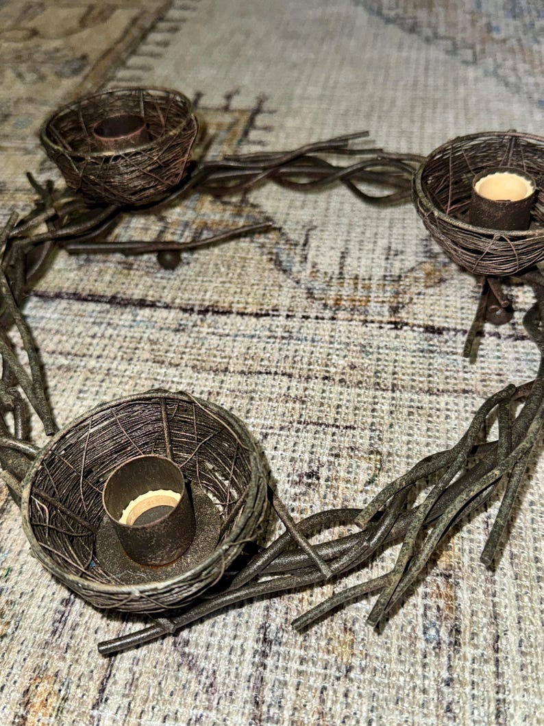 May include: A rustic metal candle holder with three candle cups shaped like bird nests. The holder is made of twisted metal branches and has a dark brown finish.