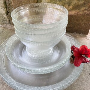May include: A stack of clear glass bowls and plates with a textured, etched design. The bowls are stacked on top of the plates, with a red flower in the background. The glassware has a decorative, textured rim.