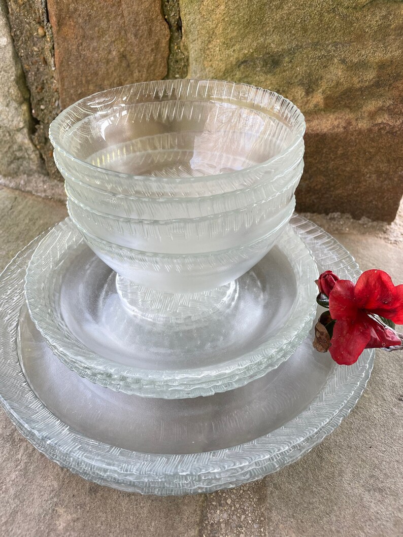 May include: A stack of clear glass bowls and plates with a textured, etched design. The bowls are stacked on top of the plates, with a red flower in the background. The glassware has a decorative, textured rim.