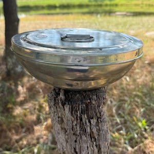 May include: A round, silver-colored metal container with a flat, circular lid. The lid features a small, centered metal handle for lifting. The container has a smooth, polished finish with visible signs of wear and a slightly tapered, bowl-like base.