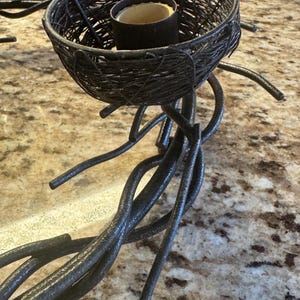 May include: A black metal candle holder with a twig design. The holder has a nest-shaped top with a small candle inside.