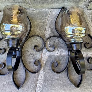 May include: A pair of wall-mounted candle sconces featuring black metal scrollwork frames and clear, amber-tinted glass hurricane shades. Each sconce has a curved metal arm supporting the glass vessel, designed to hold a candle securely in place.