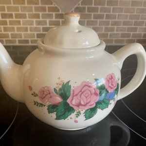 May include: White ceramic teapot with a floral design featuring pink roses and green leaves. The teapot has a lid with a small hole in the top.