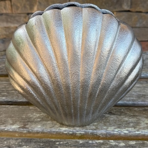 May include: A metallic silver decorative object shaped like a large scallop shell. The piece features a textured surface with prominent vertical ridges radiating from the base to the scalloped top edge, creating a fan-like appearance with a sheen.