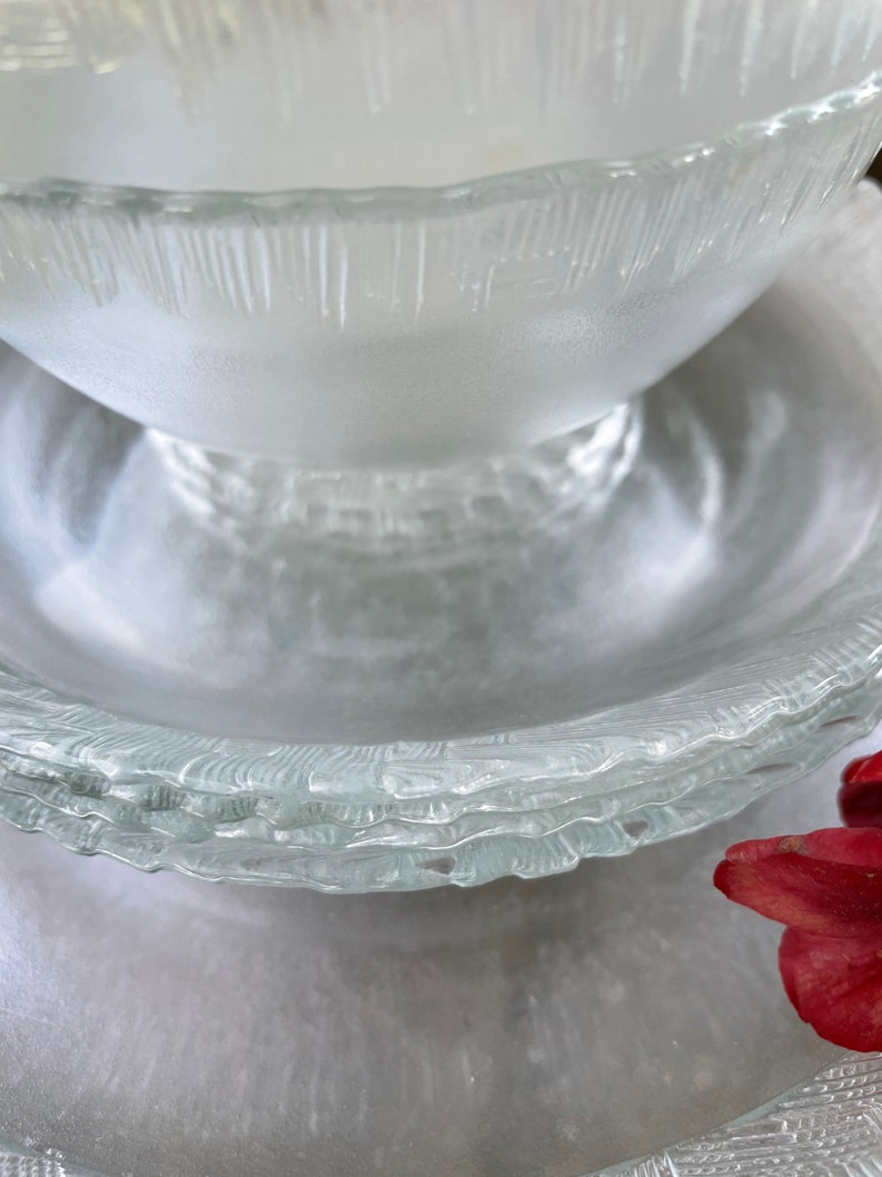 May include: A stack of clear glass bowls and plates with a textured, frosted appearance. The bowls have a rounded shape, and the plates have a wavy, textured rim. The glassware is arranged on a light-colored surface.
