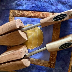 May include: Two wooden shoe trees with metal rods and leather pull loops. Each tree has a label that reads "Unmistakably Johnson & Murphy". The shoe trees are resting on a blue and gold patterned surface.
