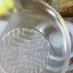 May include: A clear glass plate with a textured surface. The plate has a woven basket pattern on the bottom and a ridged edge. The plate is round and appears to be vintage.