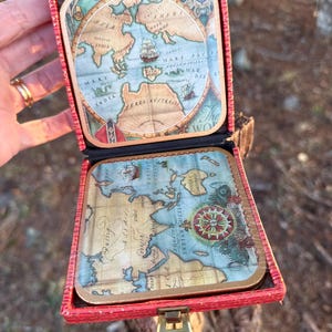May include: A set of four square coasters with a vintage map design, presented in a red box. The coasters feature detailed illustrations of continents, oceans, and sailing ships. The box has a gold-colored clasp.