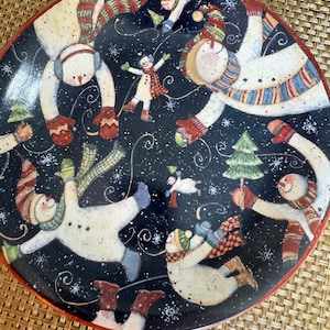 May include: A circular ceramic plate with a dark blue backdrop, decorated with playful snowmen in diverse positions. The snowmen are adorned with vibrant scarves, hats, and mittens. The plate has a red border and snowflakes.