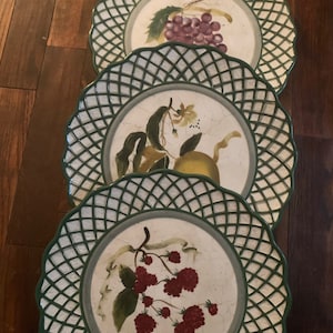 Puede incluir: Tres platos de cena de cerámica con borde de rejilla verde y centro blanco. Cada plato presenta un diseño de fruta diferente: uvas, un limón y frambuesas.
