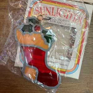 Pode incluir: Uma meia de vidro colorido vermelha e laranja com um ramo de azevinho verde. A meia está embalada em um saco de plástico com um cartão que diz "Sunlights Magical Islands of Color and Light".