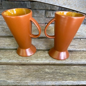 May include: Two brown ceramic mugs with a yellow glaze inside. The mugs have a flared shape and a handle on each side.