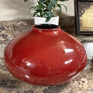 Può includere: Un vaso rotondo in ceramica rossa con finitura lucida e interno nero. Il vaso ha una forma ampia e bulbosa e poggia su un piano di lavoro maculato. Una piccola pianta in vaso è visibile sullo sfondo.