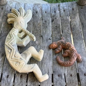 May include: A light-colored stone carving of a flute player with a zigzag pattern on the body and a smaller, reddish-brown carved lizard figure with textured details, both displayed as separate decorative objects on a wooden surface.