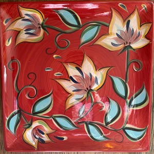 Puede incluir: Plato de servir cuadrado de cerámica con fondo rojo y diseño floral. Las flores son blancas con detalles amarillos, rosas y azules. Las hojas son verdes con detalles azules.