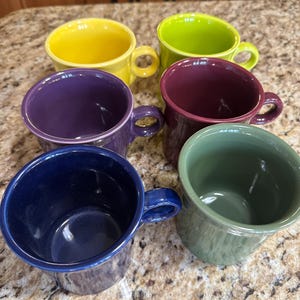 May include: Six ceramic mugs in various colors: yellow, green, purple, burgundy, navy blue, and olive green. Each mug has a rounded shape with a handle. The mugs are arranged on a speckled countertop.