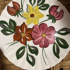 Puede incluir: Plato de cerámica blanca con borde festoneado, pintado a mano con flores coloridas. Las flores son en tonos amarillos, marrones y rosas, con hojas verdes. El plato está sobre una superficie tejida.