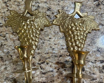 May include: A pair of gold-toned brass candle holders with a grapevine design. Each holder features a single candle cup and a curved arm for hanging.